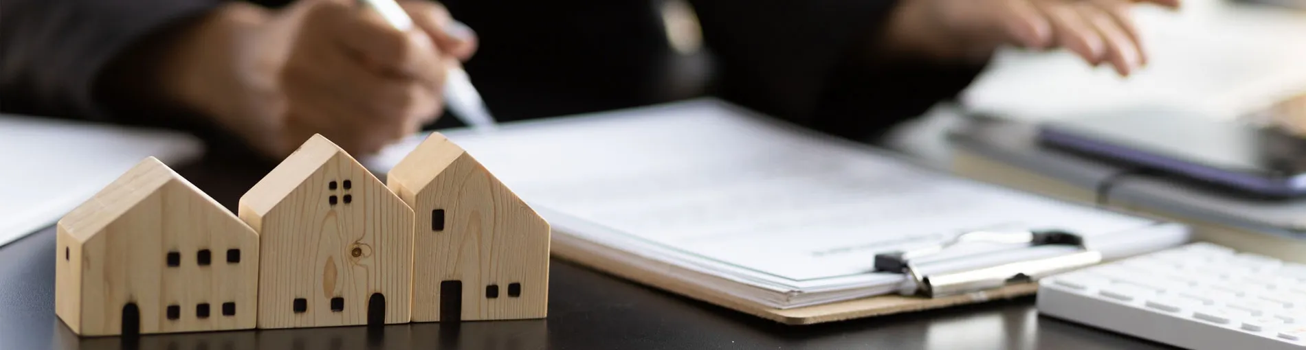 Maisons miniatures en bois posées sur un bureau, avec des documents et des mains écrivant en arrière-plan