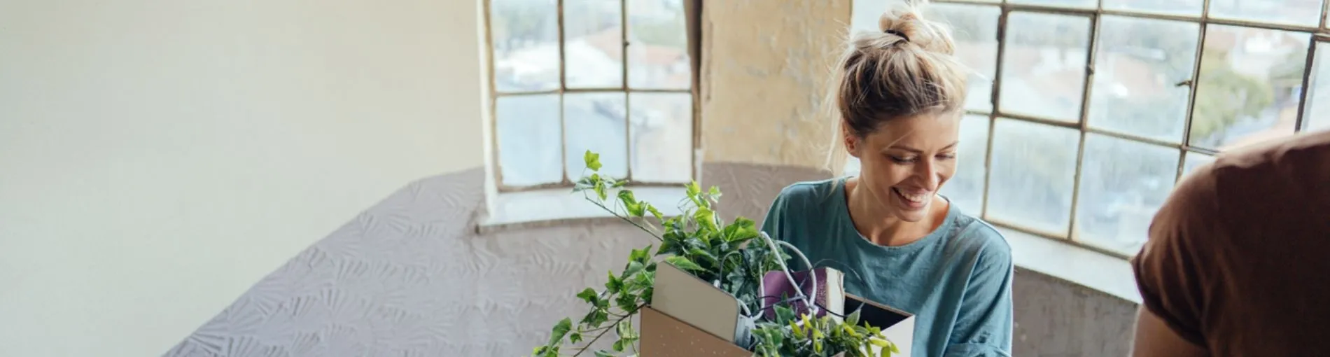 Une personne à l’intérieur d’un espace lumineux, tenant une boîte en carton remplie de plantes vertes, avec de grandes fenêtres en arrière-plan.
