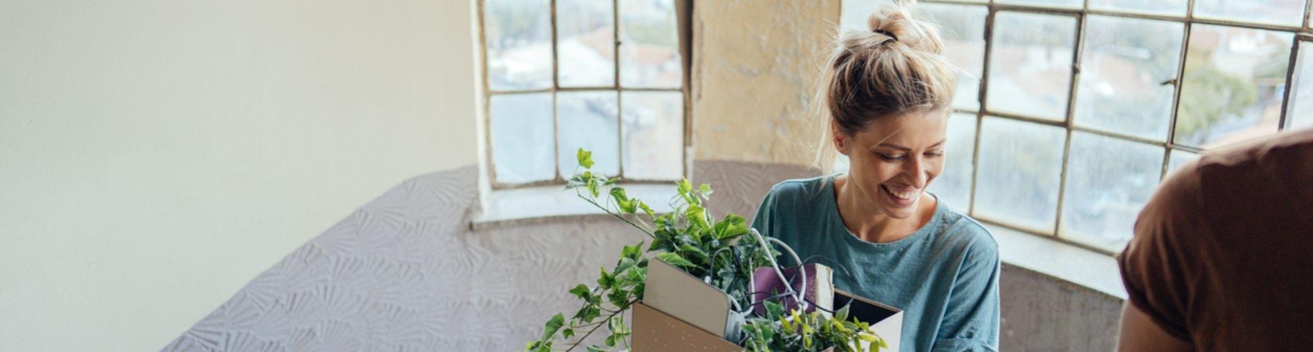 Une personne à l’intérieur d’un espace lumineux, tenant une boîte en carton remplie de plantes vertes, avec de grandes fenêtres en arrière-plan.