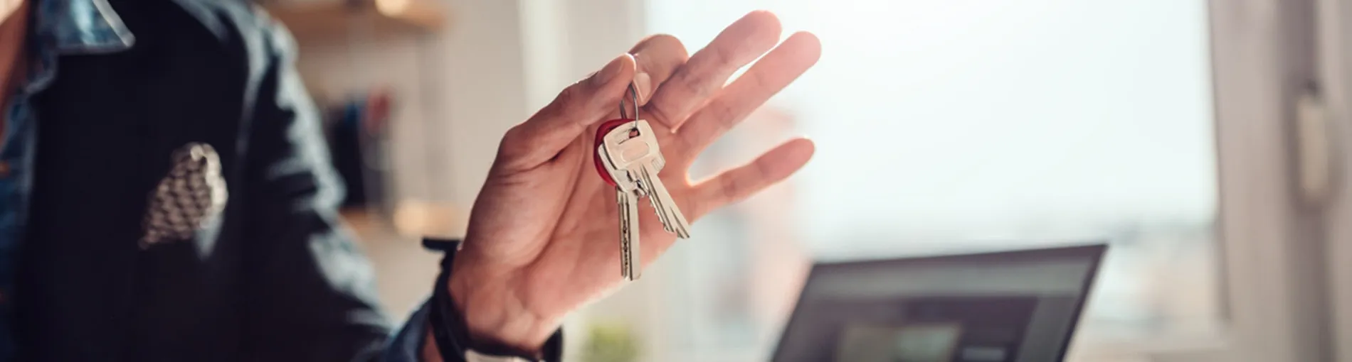 Une main tendue tenant un trousseau de clés, avec un ordinateur portable ouvert en arrière‑plan.