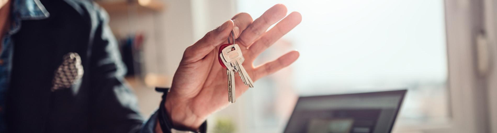 Une main tendue tenant un trousseau de clés, avec un ordinateur portable ouvert en arrière‑plan.
