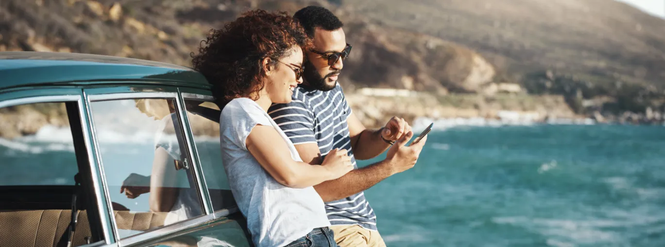 Deux personnes appuyées contre une voiture garée près de la mer, consultant un smartphone avec un paysage côtier en arrière-plan.