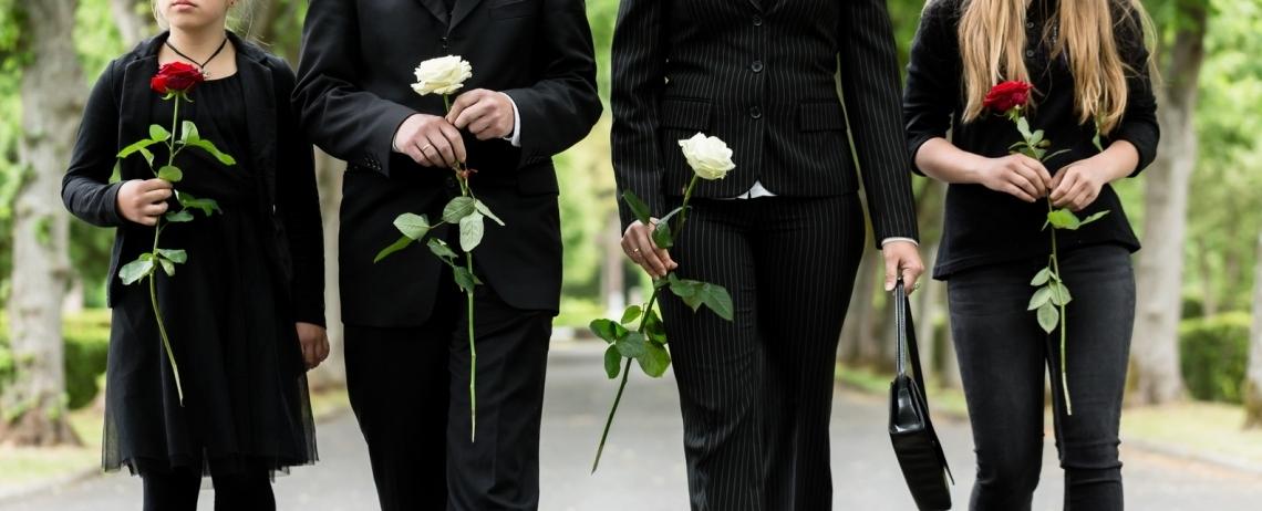 Quatre personnes vêtues de noir, vues de la poitrine aux jambes, tiennent chacune une rose à la main et marchent sur une allée bordée d’arbres, dans un cadre extérieur solennel.