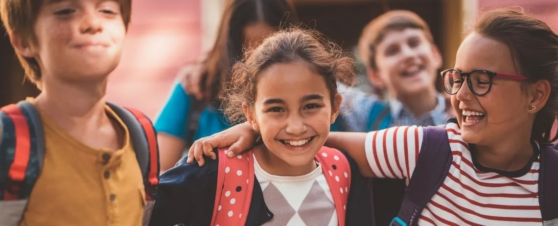 Un groupe d’enfants portant des sacs à dos se tiennent côte à côte et marchent ensemble dans un espace extérieur, devant un bâtiment, dans un contexte scolaire.