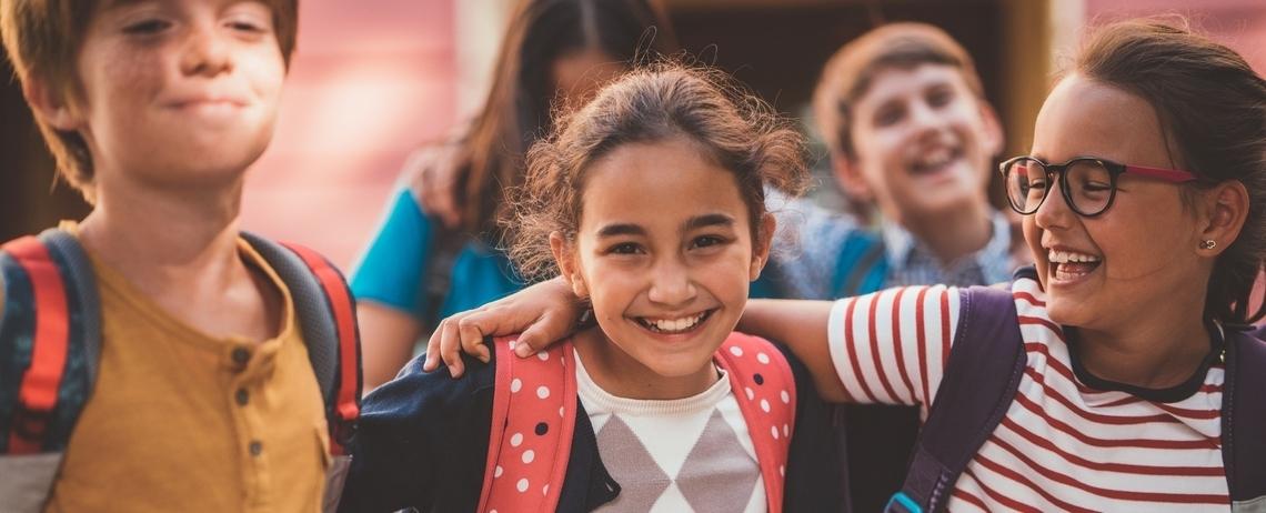 Un groupe d’enfants portant des sacs à dos se tiennent côte à côte et marchent ensemble dans un espace extérieur, devant un bâtiment, dans un contexte scolaire.