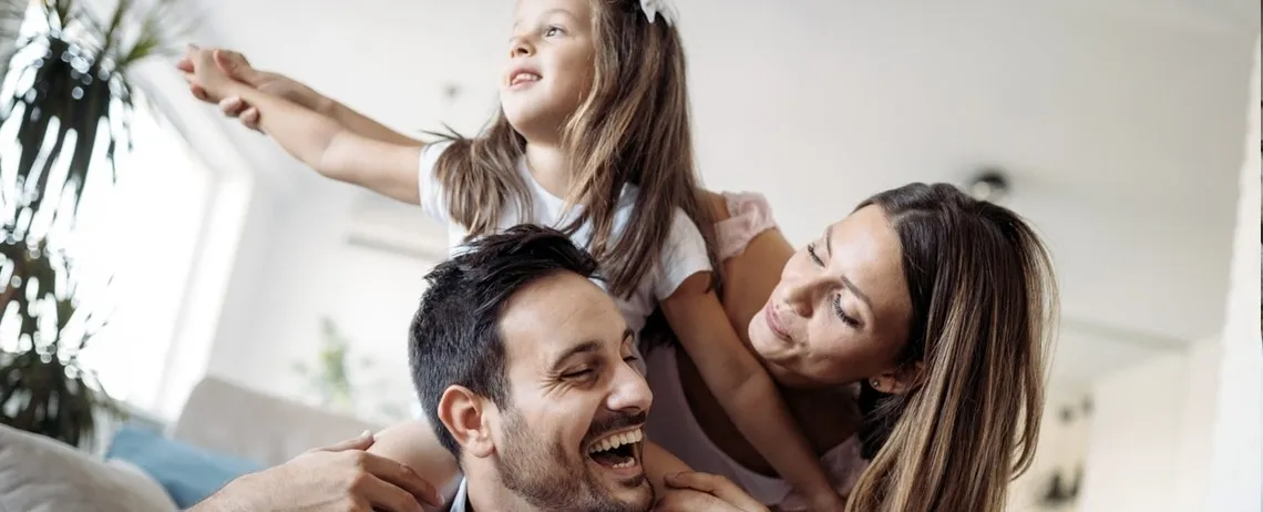 Deux adultes et un enfant réunis dans un intérieur lumineux, l’enfant porté sur les épaules d’un adulte.