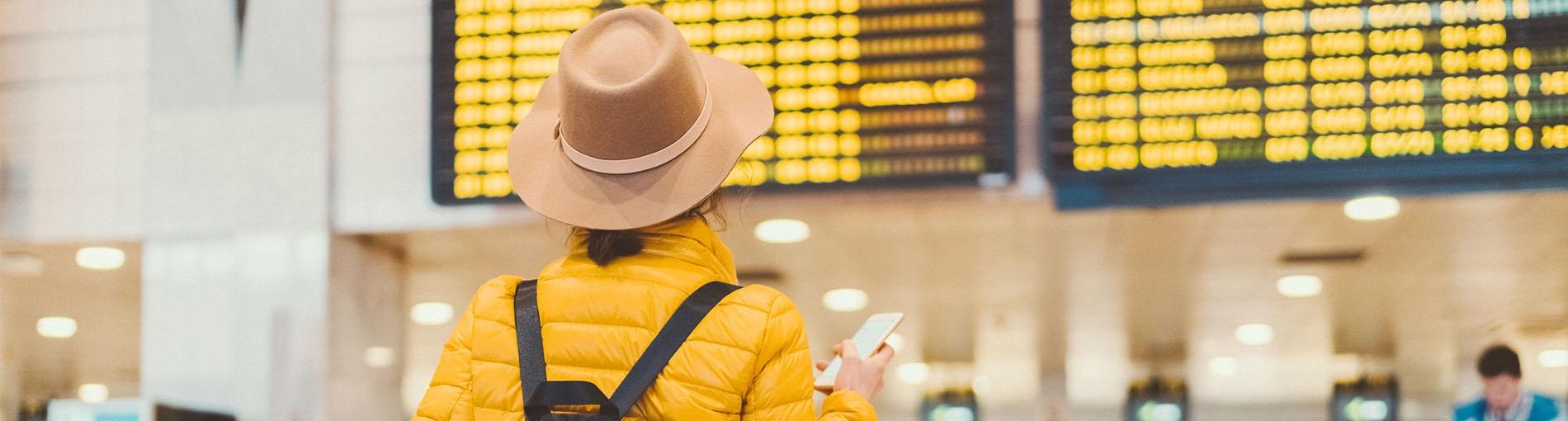 Femme de dos avec un chapeau et une doudoune jaune regardant le tableau des départs de vols dans un aéroport