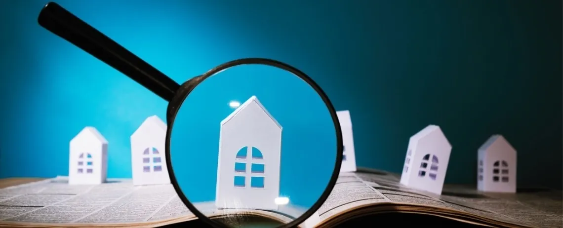 Illustration d'une loupe posée sur un livre ouvert met en évidence une petite maison miniature blanche, entourée d’autres maisons similaires, sur un fond bleu.
