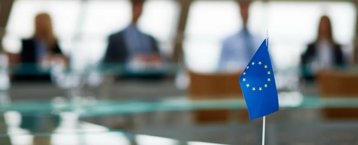 Drapeau de l’Union européenne devant une table de réunion, participants flous en arrière-plan.