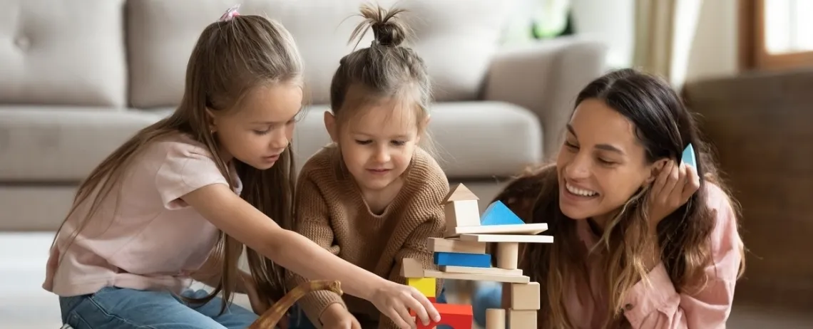 Deux enfants et un adulte sont assis au sol dans un salon, en train de construire une tour avec des blocs en bois colorés.