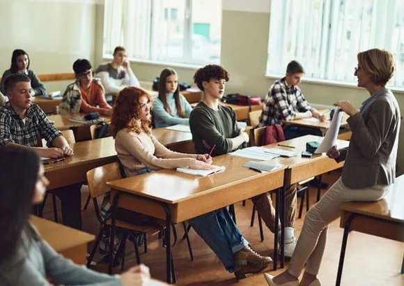 Salle de classe avec plusieurs personnes assises à des bureaux et une personne debout présentant des documents à l’avant