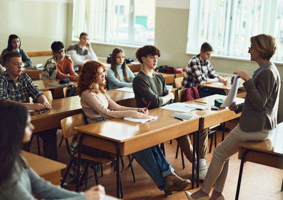 Salle de classe avec plusieurs personnes assises à des bureaux et une personne debout présentant des documents à l’avant