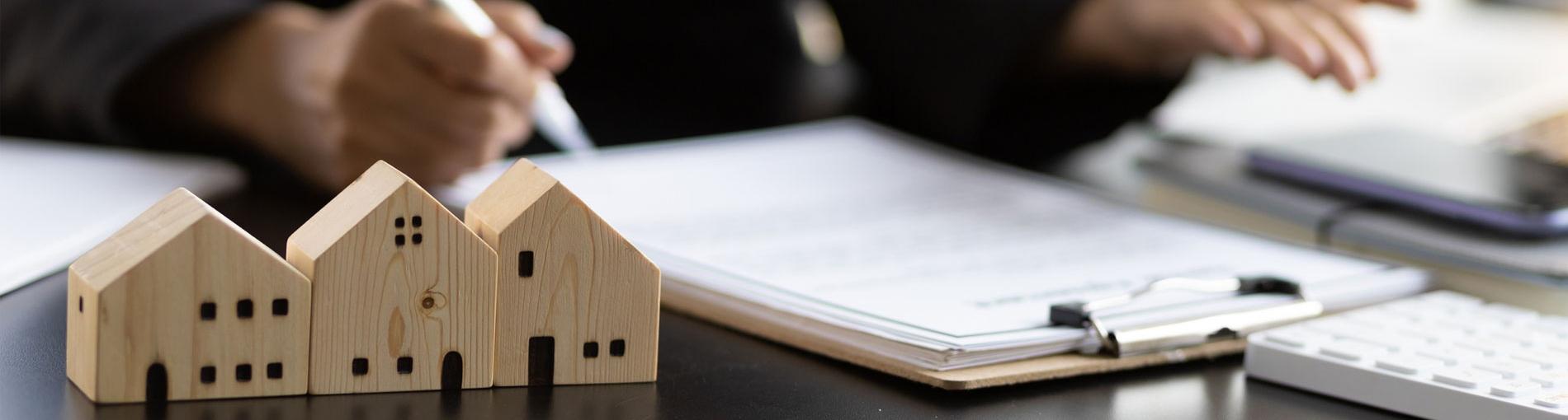 Maisons miniatures en bois posées sur un bureau, avec des documents et des mains écrivant en arrière-plan