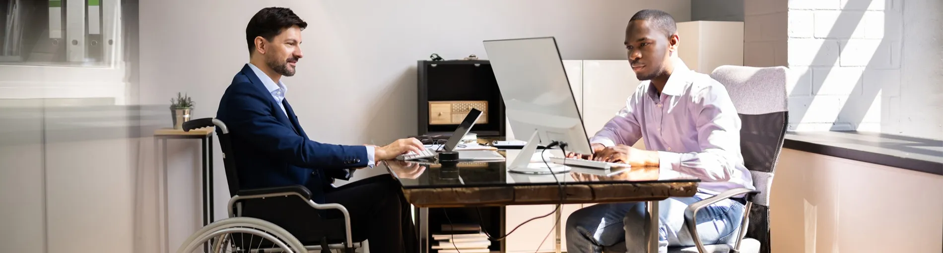 Deux personnes travaillant ensemble dans un bureau lumineux, assises à un bureau avec des ordinateurs, dont l’une en fauteuil roulant