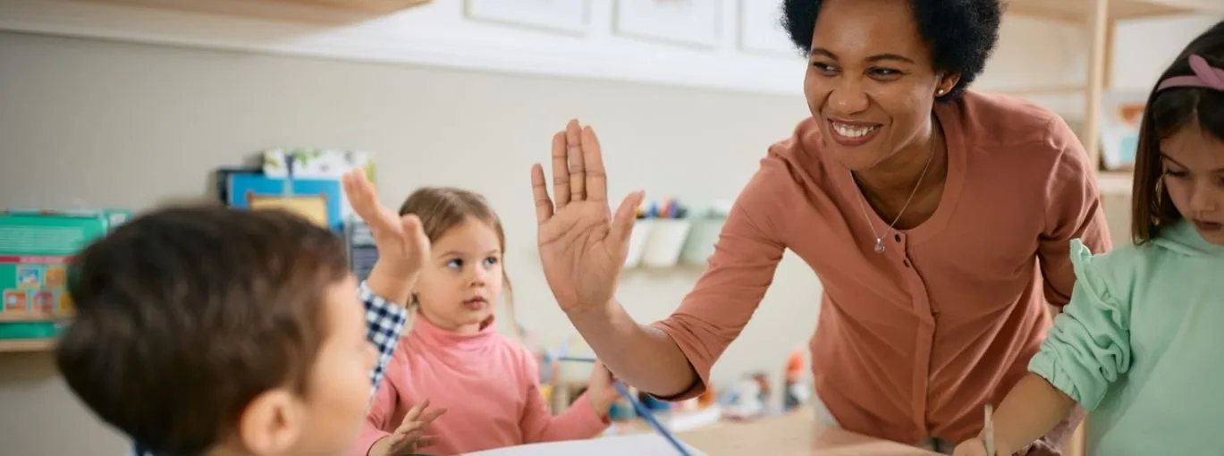Un adulte et des enfants autour d’une table, échangeant un geste de la main dans une salle d’activités.