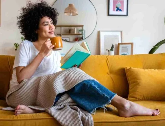 Personne assise sur un canapé jaune, tenant une tasse et un livre dans un salon.