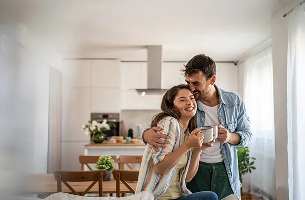 Deux adultes debout dans une cuisine lumineuse, se tenant l’un contre l’autre, l’un tenant une tasse, avec une table et des chaises en arrière‑plan.