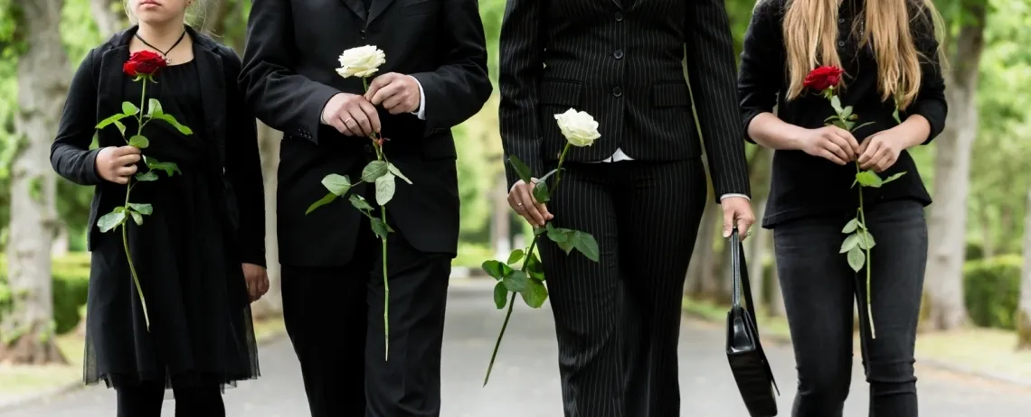 Quatre personnes vêtues de noir, vues de la poitrine aux jambes, tiennent chacune une rose à la main et marchent sur une allée bordée d’arbres, dans un cadre extérieur solennel.