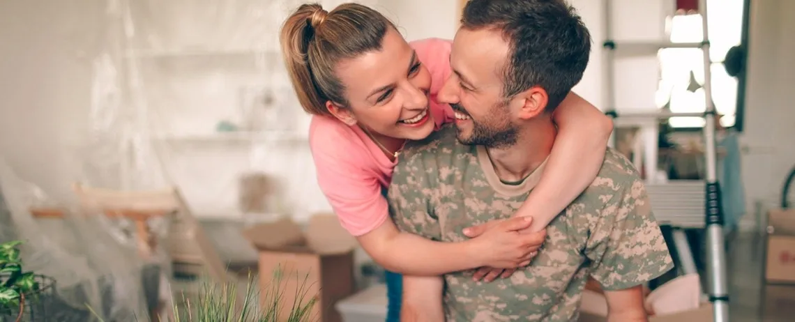 Une femme enlace un homme en habit de militaire dans un intérieur en cours d’emménagement, avec des cartons visibles en arrière-plan.