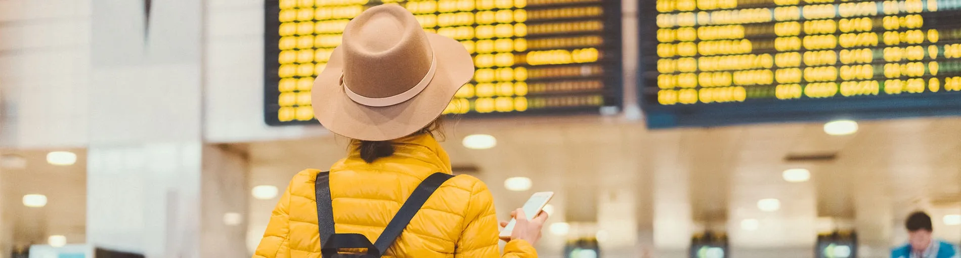 Femme de dos avec un chapeau et une doudoune jaune regardant le tableau des départs de vols dans un aéroport