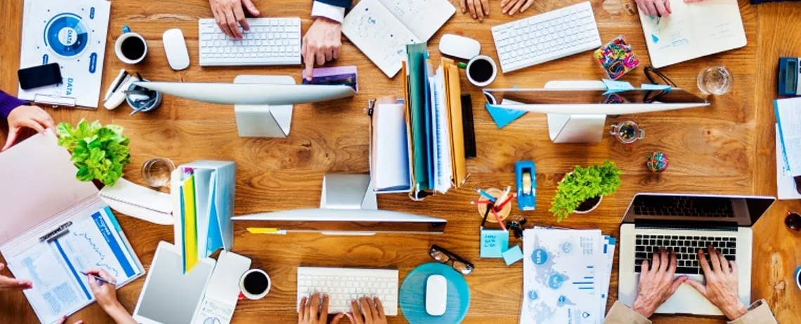 bureau vu de dessus avec beaucoup de documents, des mains posées sur un ordinateur portable, des petites plantes, des cafés