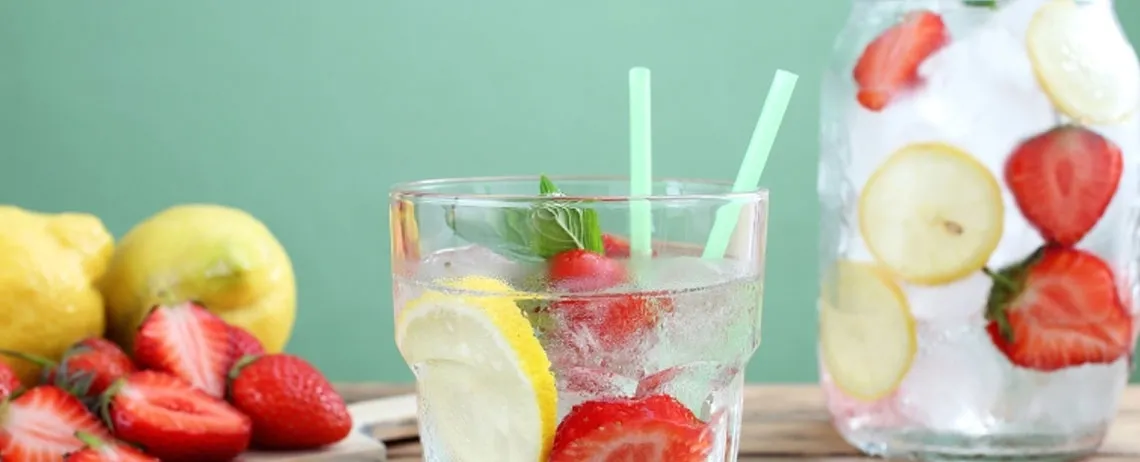 Verre rempli de boisson avec des tranches de fraise et de citron, posé sur une table, avec des fruits et une carafe en arrière-plan.